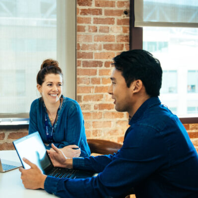 Two people sitting at a table in a meeting, engaged in conversation. One is using a laptop; both are smiling. The setting has a brick wall.