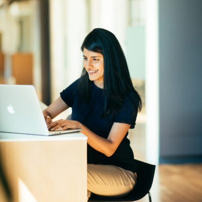 A person with long hair is smiling and typing on a laptop at a desk, representing work or study in a bright indoor setting.